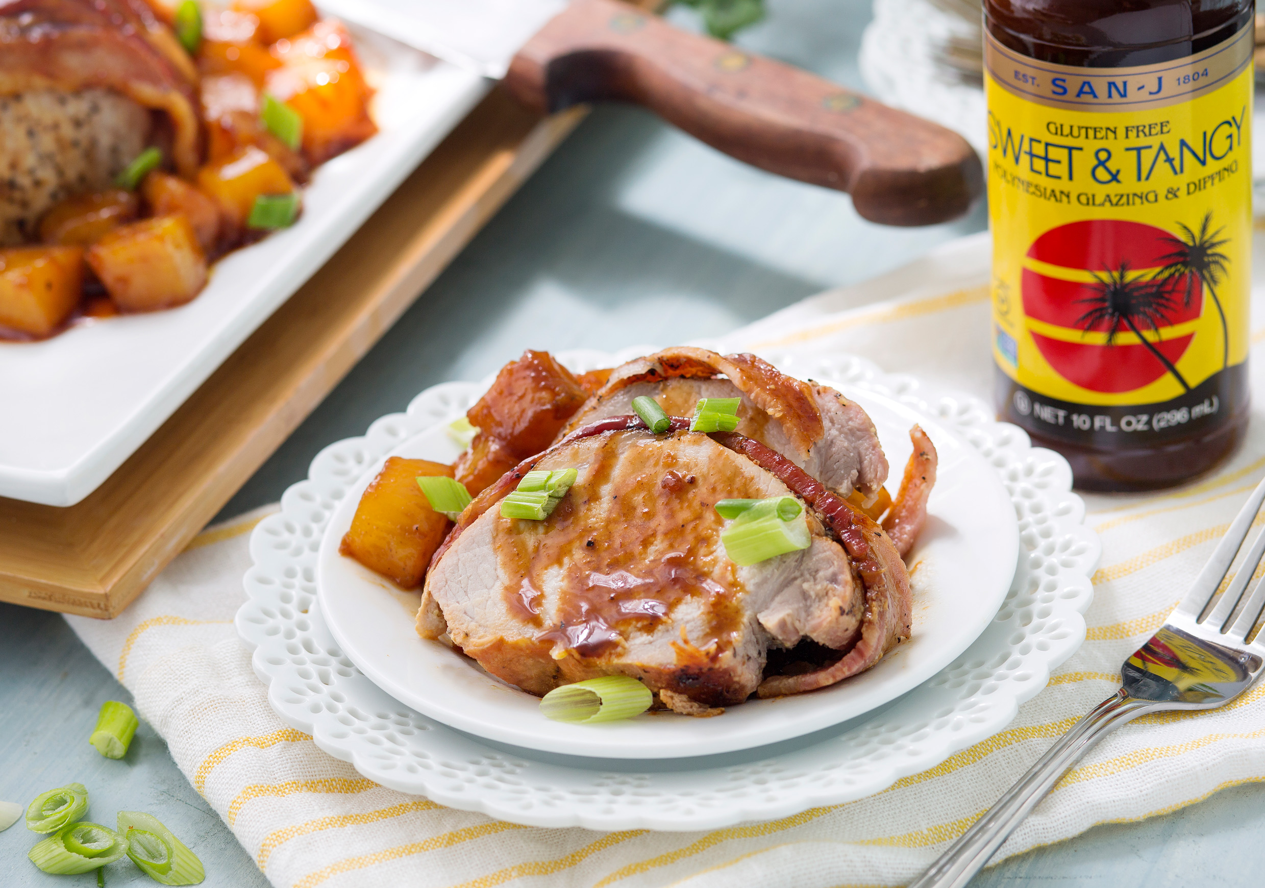 A spread on a table with bacon and pineapple pork roast next to a bottle of San-J Sweet & Tangy soy sauce